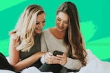 Two women scrolling through a mobile phone in bed for a story about what to do before buying a home with your partner.