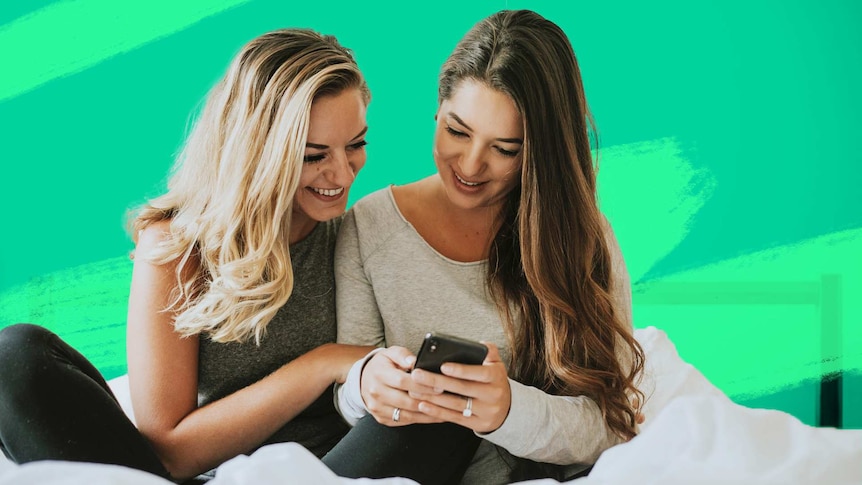 Two women scrolling through a mobile phone in bed for a story about what to do before buying a home with your partner.