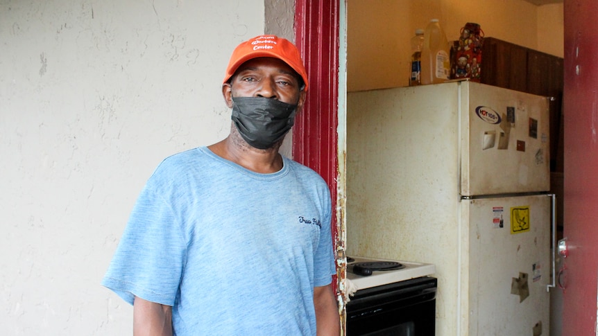 A Black man in a covid mask stands near an open door revealing a dirty fridge