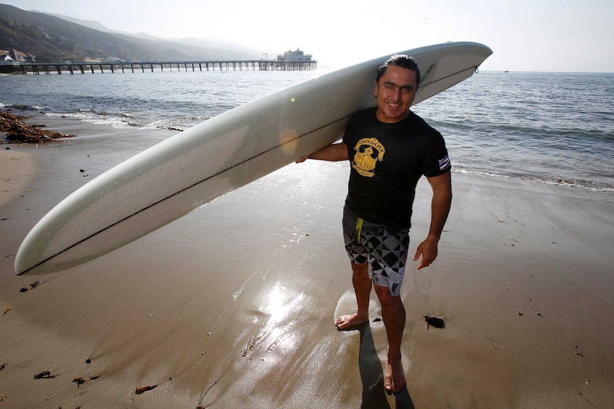 Ross Clarke-Jones stands on the beach holding his surfboard.