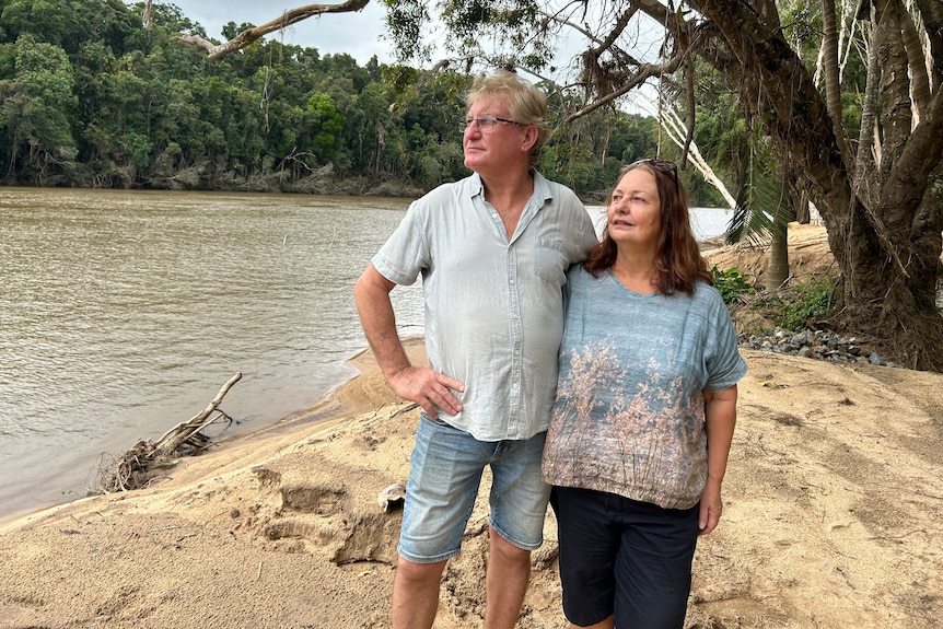 Man in blue shirt, denim shots, woman in dark shorts, printed top, stand arm in arm on a riverbank, looking away seriously.