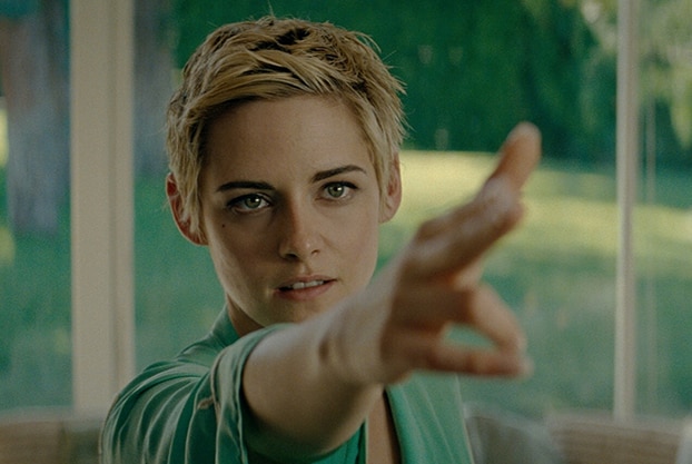 A woman with short blonde and brown hair stands in room near large window with right hand raised in gun-like gesture.