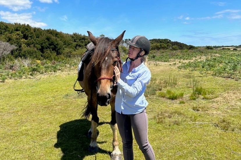 Woman wearing horse-riding helmet stands on lush green field as she caresses a horse's face. 