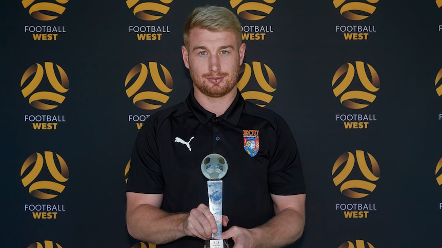 A man in an ECU Joondalup polo shirt holding a trophy, in front of a Football West background.