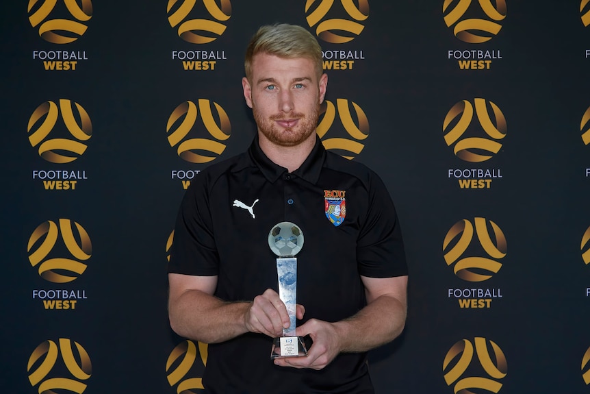 A man in an ECU Joondalup polo shirt holding a trophy, in front of a Football West background.