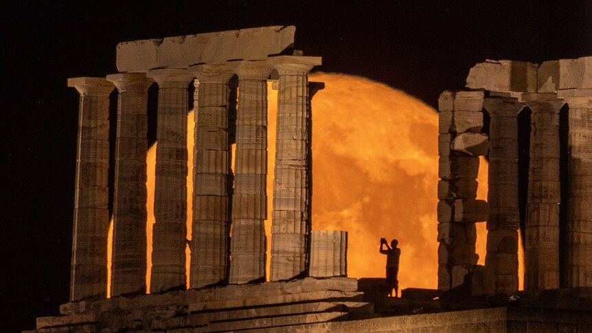 A person takes a photo as a full moon known as the "Buck Moon" rises behind the Temple of Poseidon, in Cape Sounion, near Athens