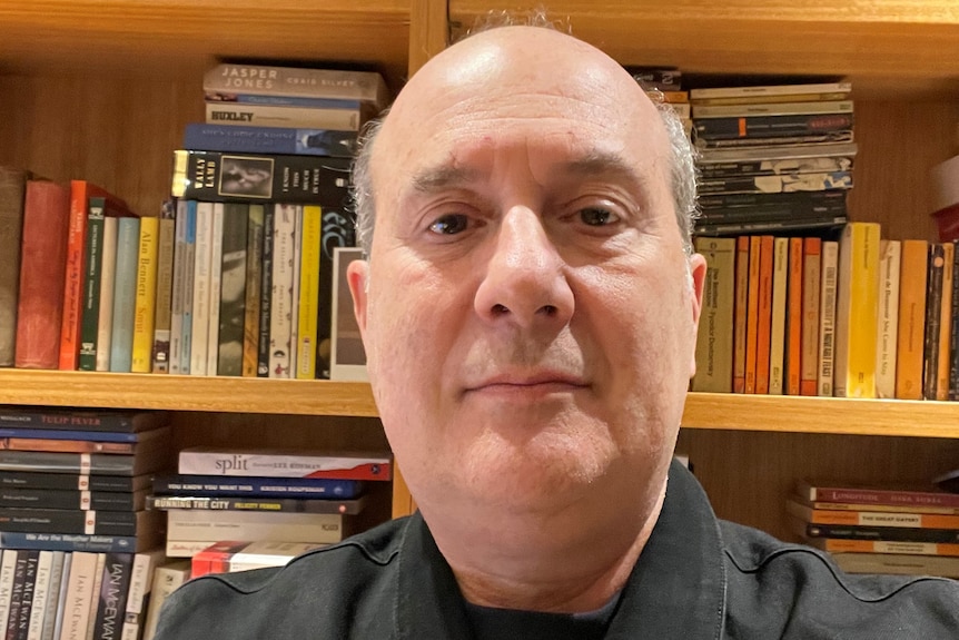 A balding middle-aged man in black looks at the camera with book shelf behind.