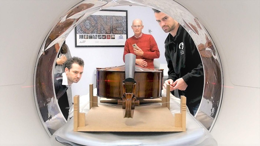 Three men peer through the aperture of a CT scanning machine, lining up a cello on the bed.