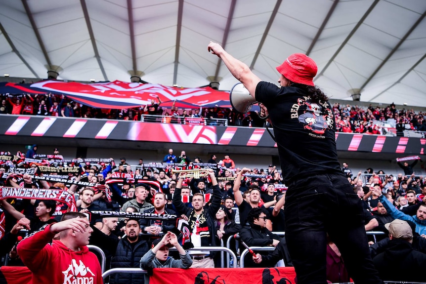 A man in a red hat and black clothing leans towards the crowd with a megaphone.