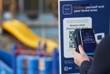 A person uses a smartphone to scan a check-in QR code outside a playground.