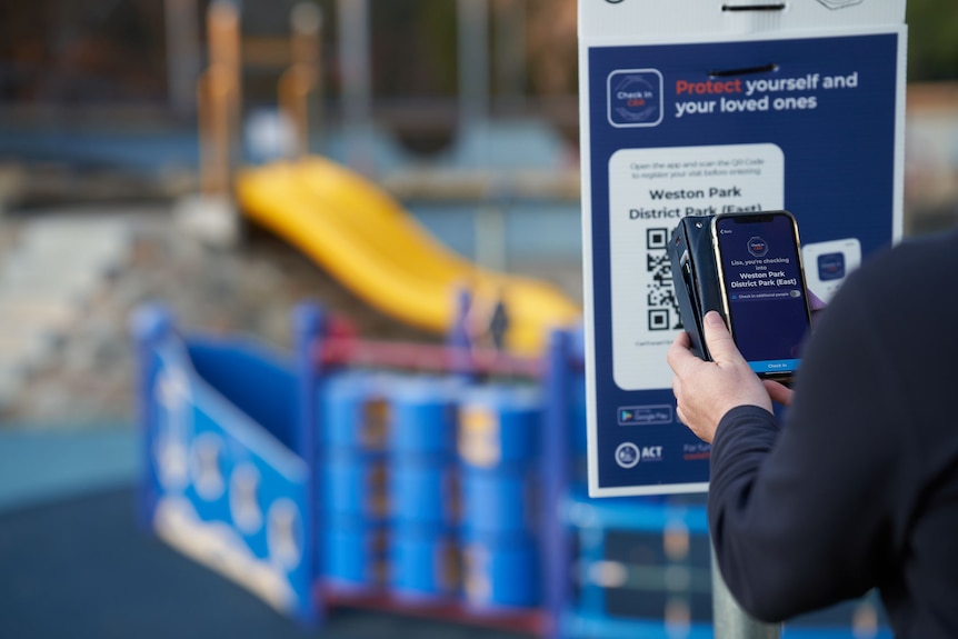 A person uses a smartphone to scan a check-in QR code outside a playground.