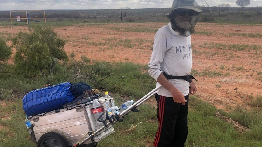 Man wearing fly net over his face walking through the outback, wheeling a suitcase behind him