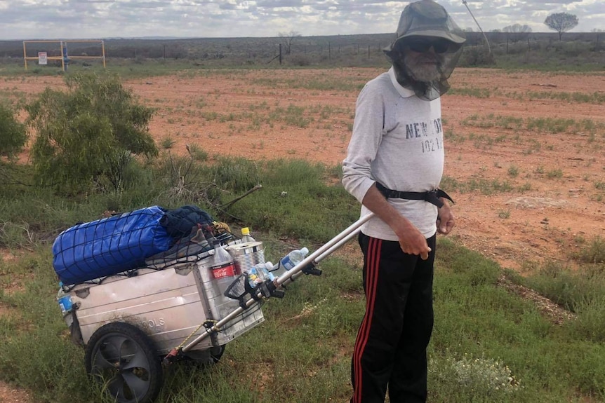 Man wearing fly net over his face walking through the outback, wheeling a suitcase behind him
