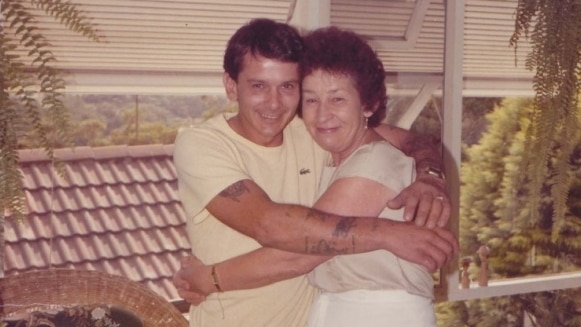 Faded colour photo of younger man and older woman hugging each other tightly and smiling widely.