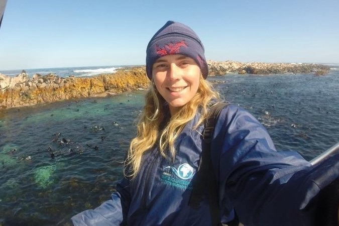 A selfie image of a young Caucasian woman by the ocean, wearing a beanie and waterproof blue jacket.