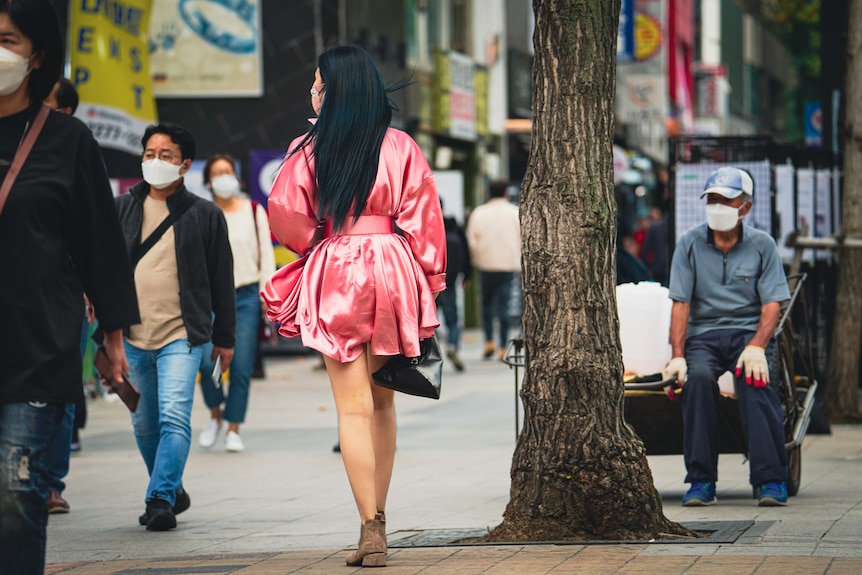 A woman in a pink dress walks down a street in Seoul, past a man sitting next to a rubbish collection wagon