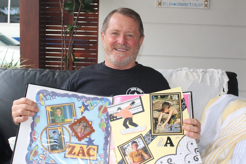 A middle aged man is sitting on a lounge, holding open a colourful scrapbook. The pages are filled with photos of a young boy