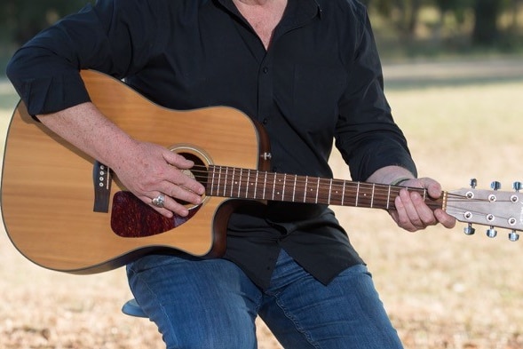 A mid-shot of an unidentified man sitting on a stool playing a guitar outside.