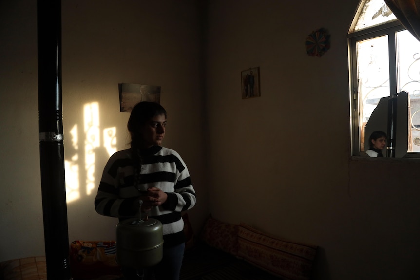 A young woman with dark braided hair wearing a holds a container in a dimly lit bedroom.
