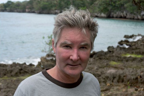 A man with thick grey hair in grey t-shirt stands before a background of water and rocks, looking to camera with slight smile.