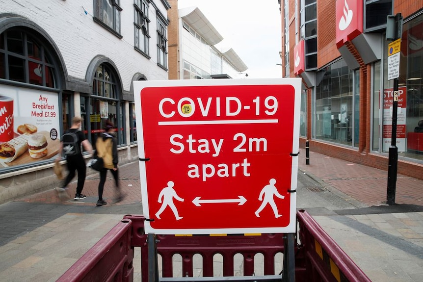 A red sign on  UK street reads COVID-19 stay 2m apart with description of two people walking socially distanced