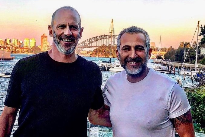 Two men stand side by side and smile at the camera with Sydney harbour at their backdrop