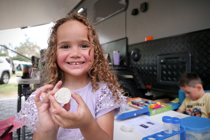 A little girl with curly hair smiles. She is showing the camera a piece of hand made art, there is a caravan in background