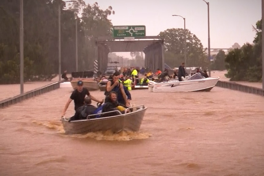 A road bridge submerged in floodwater with boats and people being rescued