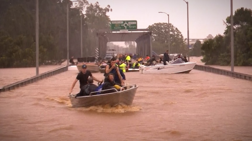 A road bridge submerged in floodwater with boats and people being rescued