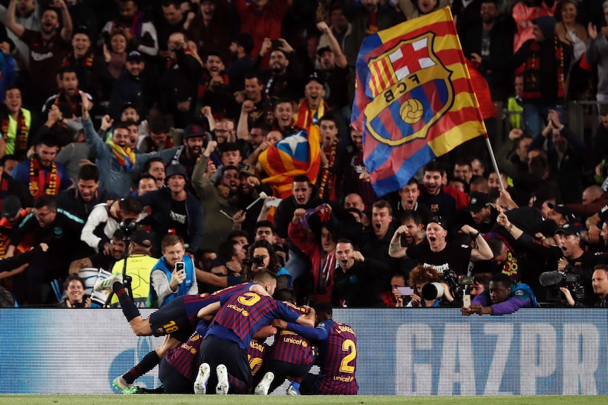 Football players pile on top of their goalscoring teammate as delirious fans celebrate in stands.