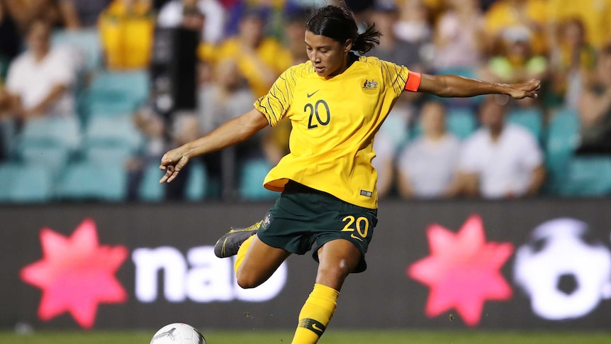 Sam Kerr wearing a yellow Matildas team shirt kicks a soccer ball on the turf of a sports arena.