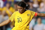 Sam Kerr wearing a yellow Matildas team shirt kicks a soccer ball on the turf of a sports arena.