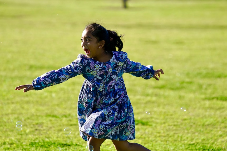 A little girl in a purple floral dress spins on the grass with a big smile on her face