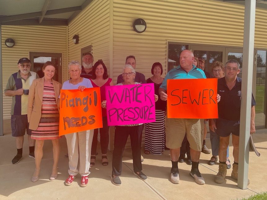 A group of adults stands under a verandah posing for a photo, with signs saying 'piangil needs', 'water pressure', and 'sewer'