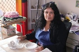 An Indigenous woman sits at a table surrounded by art supplies with ceramic items in front of her.