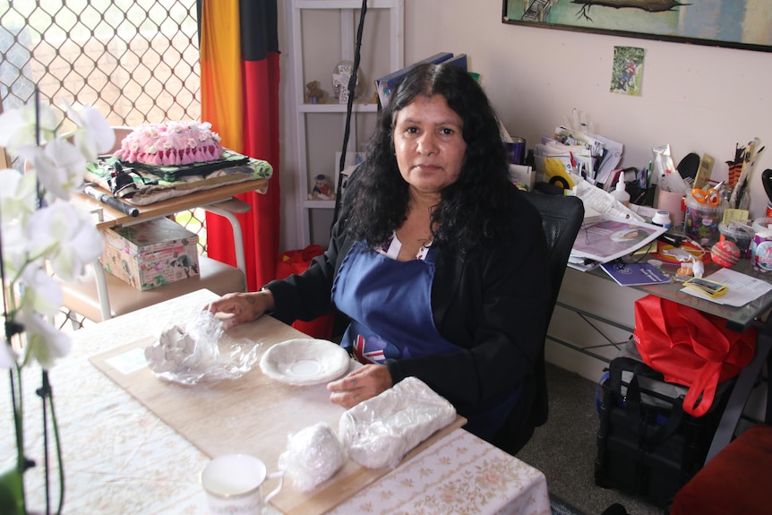 An Indigenous woman sits at a table surrounded by art supplies with ceramic items in front of her.