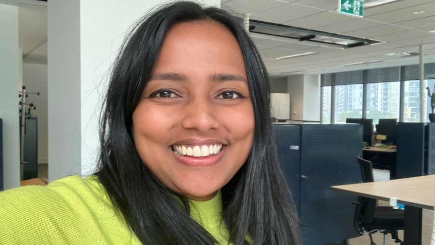 A woman with black hair and a green top smiles at the camera, with some filing cabinets and a desk behind her.