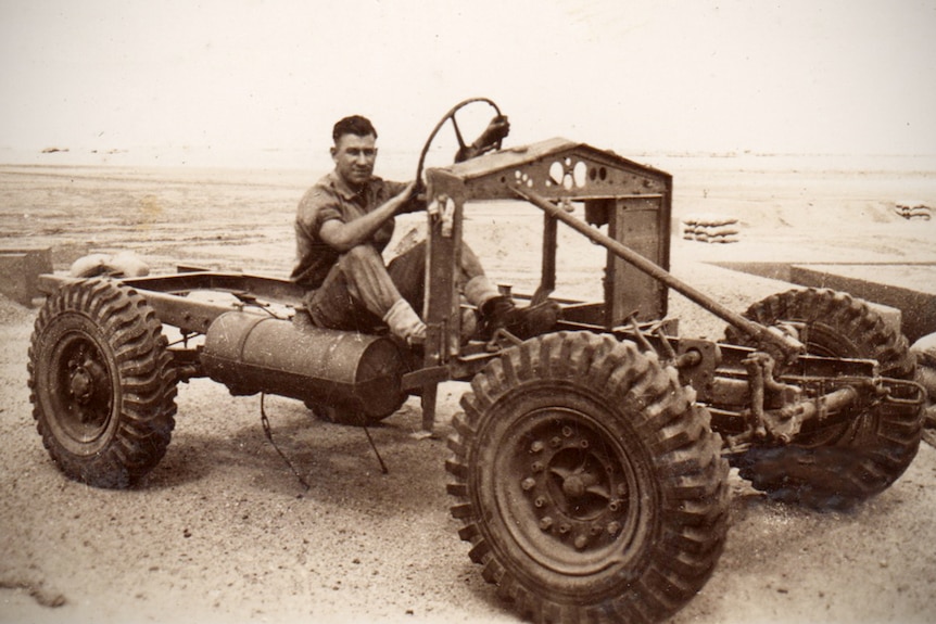 Man in work clothes sits in the metal frame of an army vehicle, holding onto steering wheel.