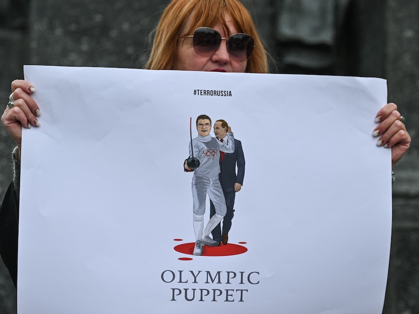 A woman holds up a sign showing Thomas Bach hugging Vladimir Putin.