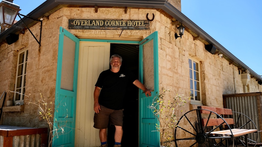 A man stands in the blue doorway of a building with a sign reading 'Overland Corner Hotel'