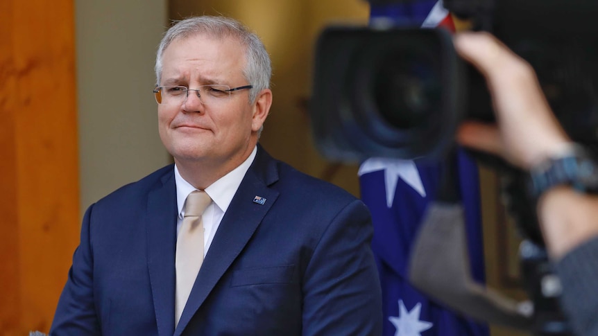 Scott Morrison looks to crowd of journalists in front of him that cannot be seen in the shot. He wears glasses and a blue suit.