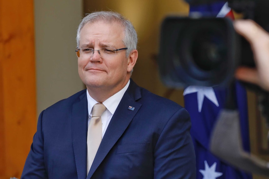 Scott Morrison looks to crowd of journalists in front of him that cannot be seen in the shot. He wears glasses and a blue suit.