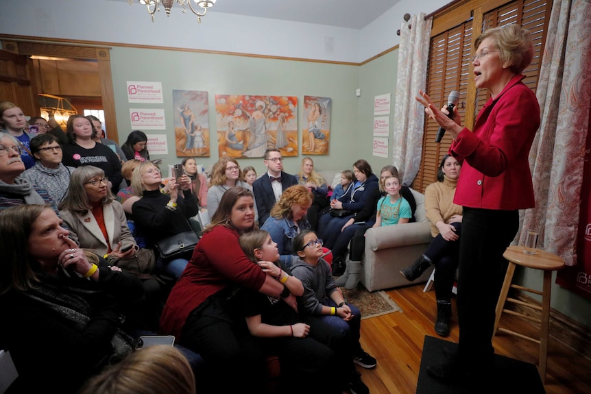 Elizabeth Warren speaking into a microphone before an audience in a living room
