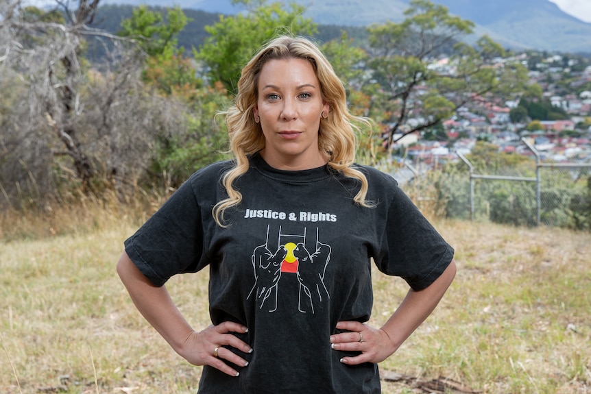 A woman poses for a photo with her hands on her hips in front of a mountain. She's wearing a t-shirt with the Aboriginal flag
