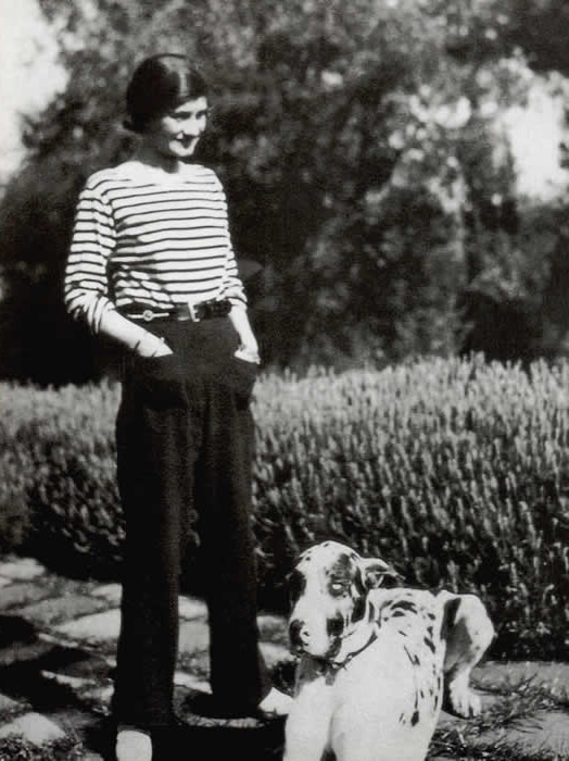 A woman in a striped top stands, hands in pant pockets, next to a dog. (Dog out of frame in some crops)