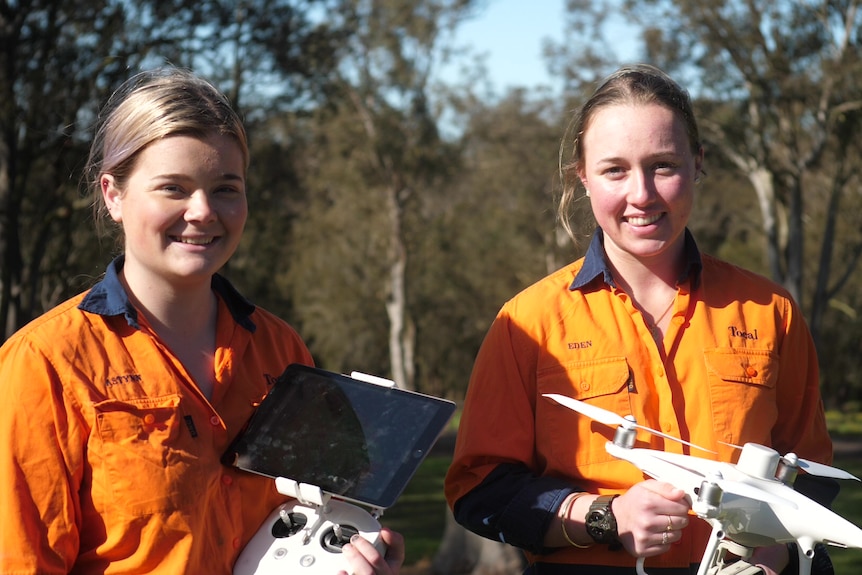 Two young students smile at the camera, student on the left is holding an ipad, student on the right is holding a drone.