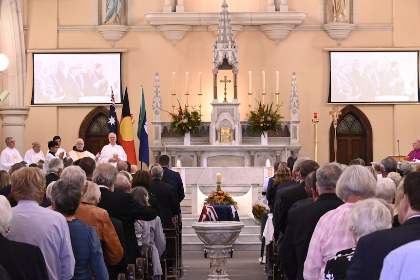 mourners seen from behind, in front of a casket draped in an australian flag