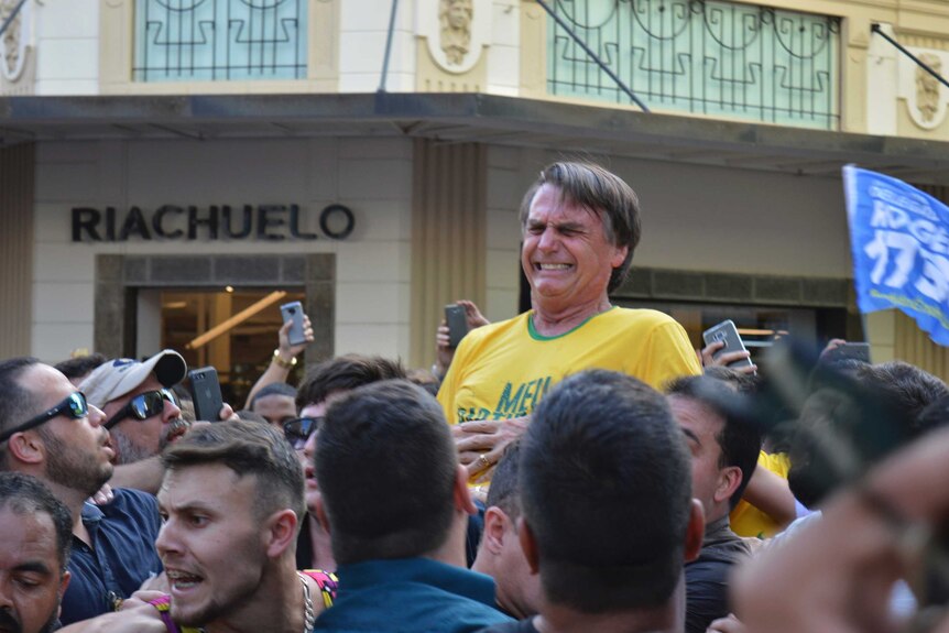 Jair Bolsonaro, who is being carried, grimaces right after being stabbed, with people all around him.