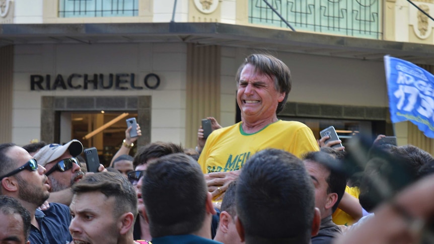 Jair Bolsonaro, who is being carried, grimaces right after being stabbed, with people all around him.