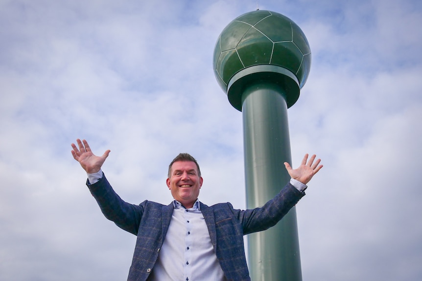 A suited man stands smiling in front of a tall, thin, green tower with a spherical top.